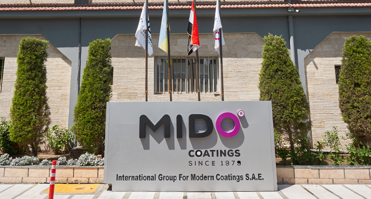 Exterior view of the MIDO Coatings facility, part of the International Group For Modern Coatings S.A.E. The building has a beige brick facade with blue accents and a sign displaying the company logo and name. Flags and neatly trimmed shrubs adorn the entrance.